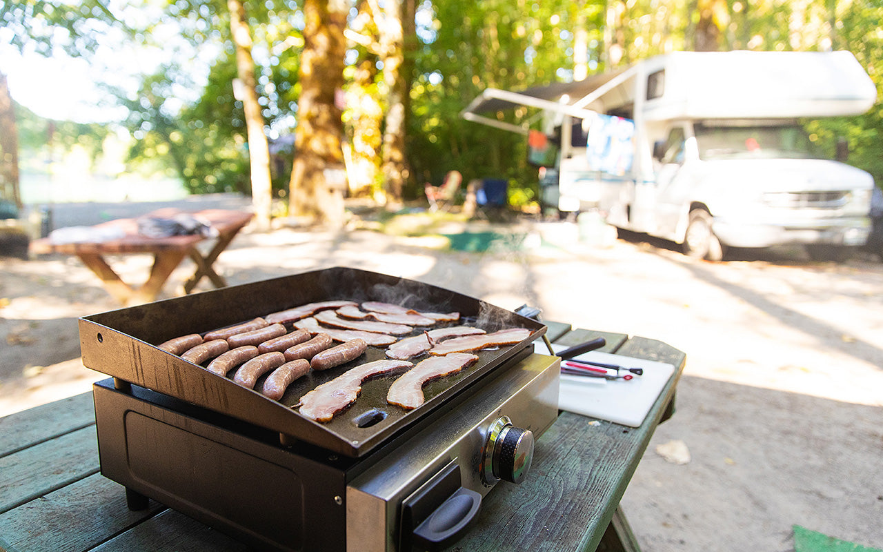 Bacon and sausage cooking on a griddle, set on top of a picnic table at a campsite with an RV in the background.